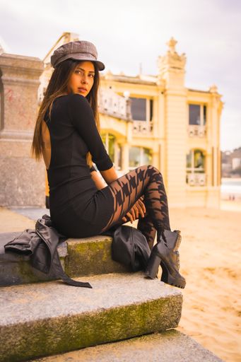 A Caucasian brunette sitting on some stairs in a short black dress, with a beret and plaid stockings next to a beach on vacation in summer