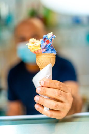 A vertical shot of a colorful ice cream cone held by a caucasian male in a mask