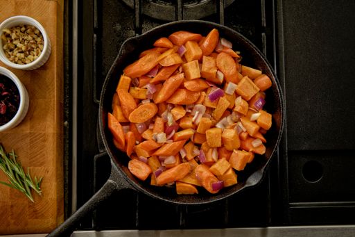 A top view of a cast iron pan with chopped carrots on a stove beside bowls of nuts
