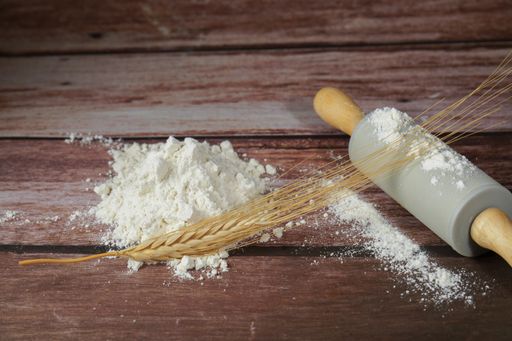 pile of flour with an ear of wheat on top of it, next to a rolling pin, on a wooden table and copy space
