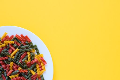Top view of red green and yellow dry raw fusilli pasta on a plate with yellow background and copy space.