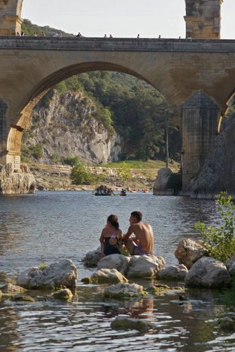 Outdoor scenic vertical summer closeup on a couple of people sitting at the riverside in front of the three-tiered aqueduct Pont du Gard in France