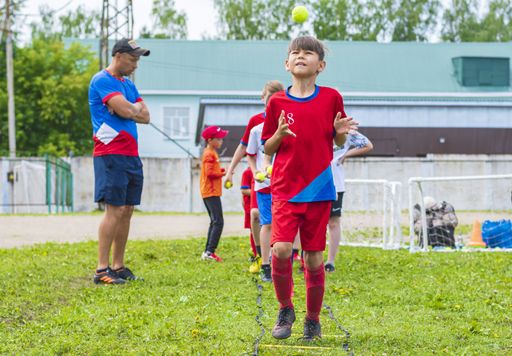 A young football player in a red uniform performs a training exercise for coordination of movement using a tennis ball. Outdoor sports.