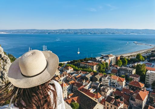 Rear view of young woman wearing stylish white summer dress and sun hat standing on lookout point over city of Omis in Croatia.