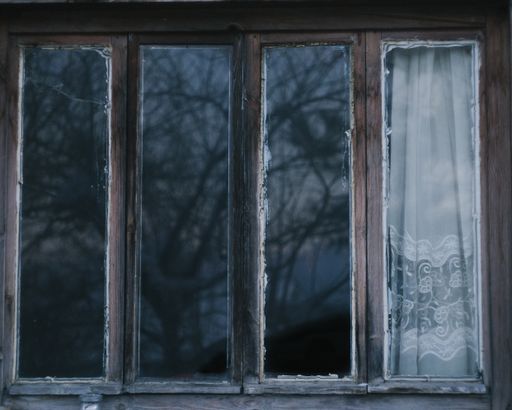 An old rusty wooden window with trees reflected on glass with white curtain.
