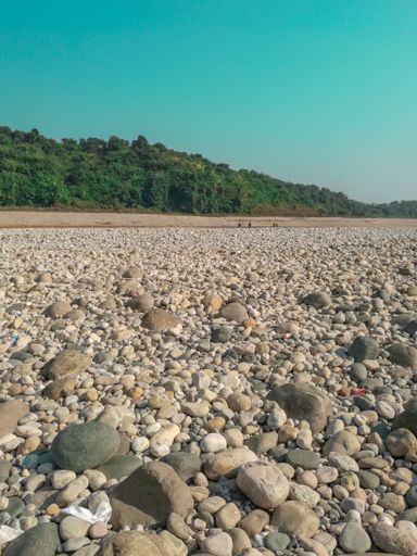 A vertical shot of a sunny day at the beach with huge rocks filling the ground and a green hill under a clear sky in the background