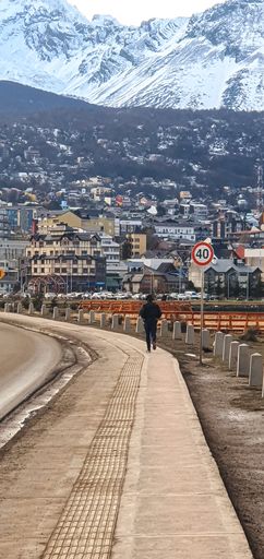A vertical shot of person walking down the roadside pavement - mountainous town on background