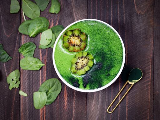 A green kiwi smoothie in a white ceramic container with a brass-colored spoon on a brown wooden surface