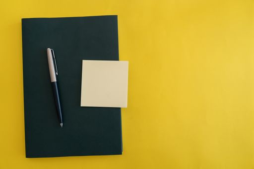 Dark green book with pen and sticky notes on yellow flat background.