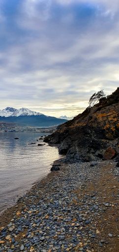 A vertical shot of a rocky shore surrounded by a lake and snowy mountains under a cloudy sky
