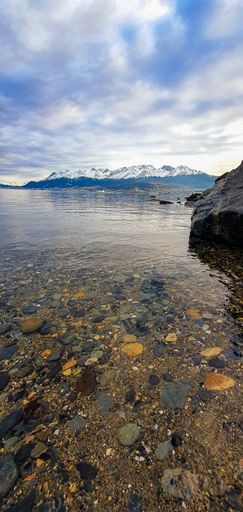 A vertical shot of a lake surrounded by rocky and snowy mountains under a cloudy sky