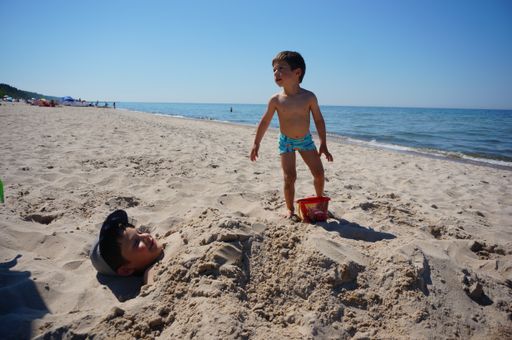Young boys digged in the sand and holding a toy bucket at a sandy beach on a warm day.