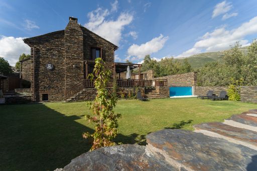 Rural house with a stone facade and a large stone patio, equipped with a swimming pool and several chairs.
