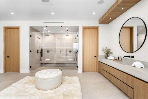 A white bathroom with a marble countertop, wooden cabinets, and a round mirror.