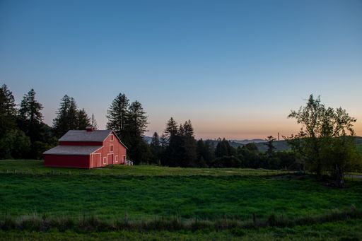 A Red barn in the middle of a green field with trees, sky and landscape in the distance