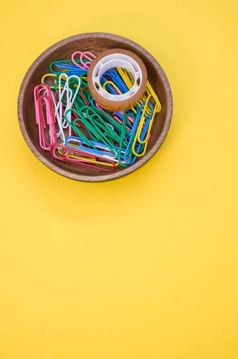 A vertical shot of colorful paper clips and an adhesive tape in a wooden bowl on a yellow background