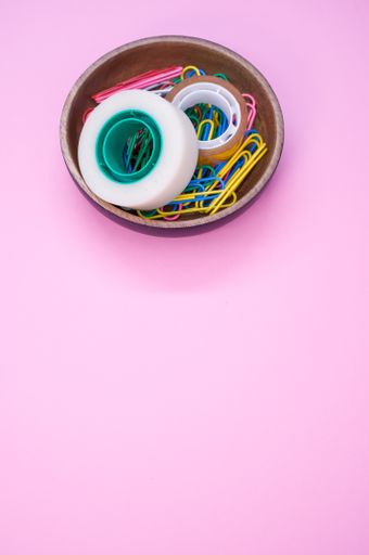 A vertical shot of colorful paper clips and adhesive tapes in a wooden bowl on a pink background