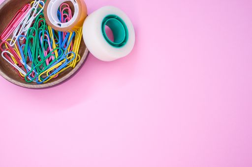 A top view of colorful paper clips and adhesive tapes in a wooden bowl on a pink background