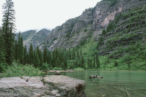 a wide shot rock near the Avalanche Lake with people walking on a shore in the distance with trees and mountain in the background