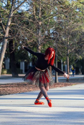 A beautiful red haired ballerina gracefully dancing at a park with trees in the background