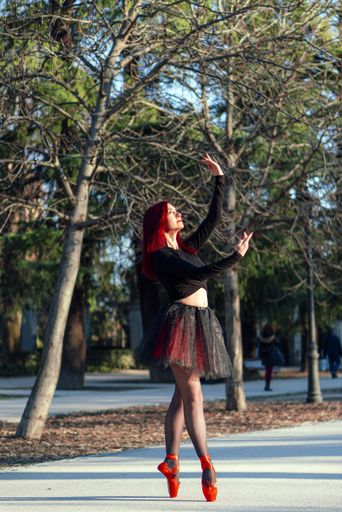 A beautiful red haired ballerina gracefully dancing at a park with trees in the background