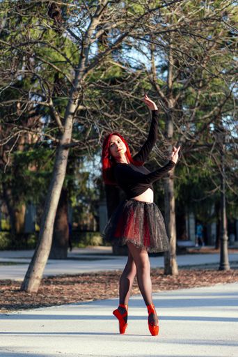 A beautiful red haired ballerina gracefully dancing at a park with trees in the background