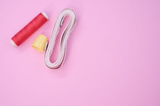 A top view of a red sewing thread with a thimble and a measuring tape on the pink surface