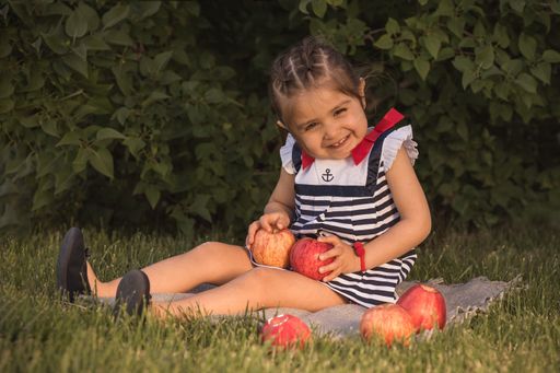 A sitting little girl holding two apples looking at the camera with green leaves in the background