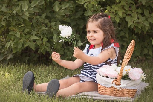 A little girl sitting near a basket holding a white flower with green leaves in the background