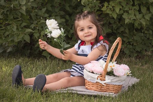 A little girl sitting near a basket holding a white flower with green leaves in the background