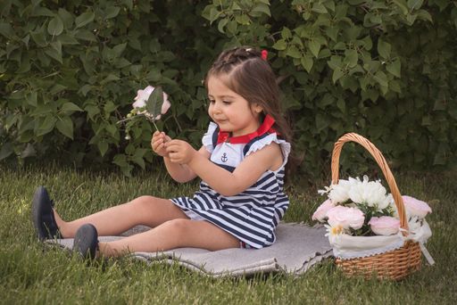 A little girl sitting near a basket holding a white flower with green leaves in the background