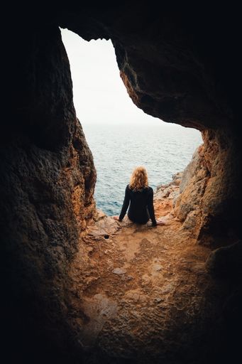 A vertical shot of a female seating in a small cave in The Balearic Islands and looking at the sea