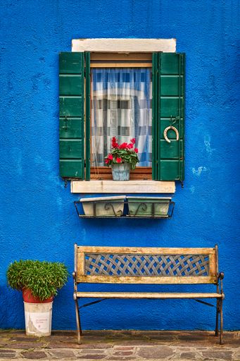 A vertical shot of a blue wall with an opened window and flowers on the sill with a bench under it