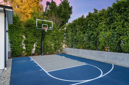 An outdoor basketball court by house with trees, bushes, and fence.