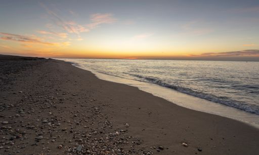 beach lies empty, beneath a sky awash in breathtaking shades of orange, its fine sands interspersed with pebbles. The sea remains tranquil.