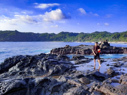 Young wet Asian man in front view standing daydreaming in a water puddle on a rock plateau with a bag of chips at Wediombo Beach, Java, Indonesia