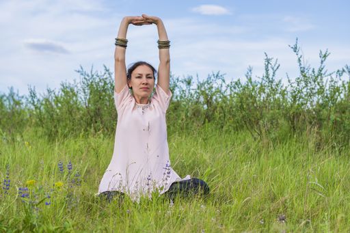 A beautiful young woman sits on a green lawn doing physical exercise with her arms high. Outdoor activities.