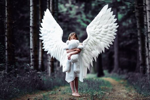 A closeup shot of a Caucasian blonde girl in angel wings costume with a teddy bear toy