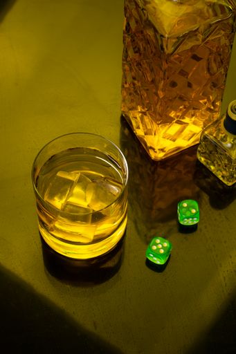 A top view of a glass of Whiskey with ice next to a bottle, green dice on glass table