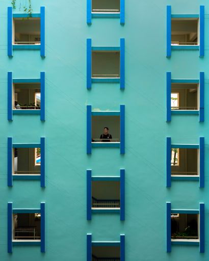 young Chinese man looking out of window of a turquoise facade. Beautiful symmetrical building exterior. Thoughtful , pensive , contemplative concepts