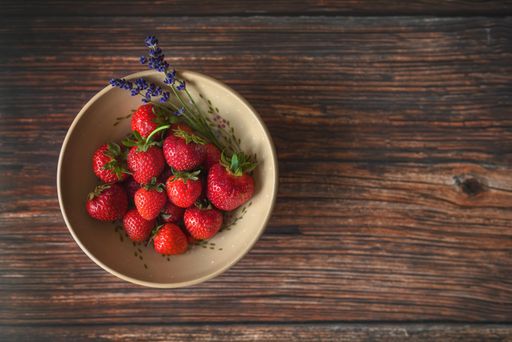 Flat lay photo of strawberries decorated with plucked lavender in a ceramic bowl lying on a wooden table.
