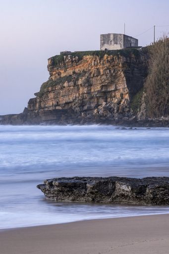 A vertical picture of a building on a cliff surrounded by the sea under a cloudy sky