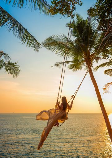 Young woman in a flowing dress swaying on a seaside swing at sunset