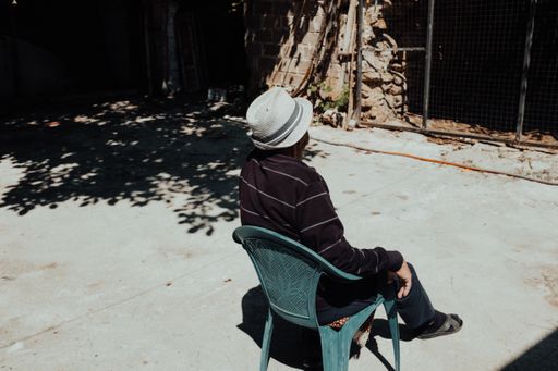 An adult person with a trilby hat sitting on a plastic chair outdoors on a sunny day