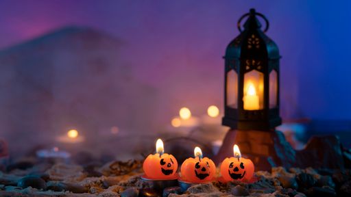Halloween background, 3 pumpkin-shaped candles in the foreground, an old lamp on a rock against a blue out-of-focus background with yellow lights