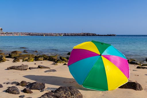 Beach umbrella of various colors, placed on the sand of a yellow sand beach near the sea with rocks around with blue sea and sky