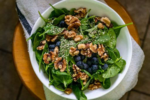 Above fresh healthy moist green spinach, blueberry, walnut and hemp hearts salad in white bowl on white towel on wooden stool.
