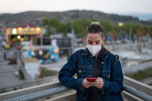 A stylish young Spanish woman wearing a facemask and using her phone on a bridge