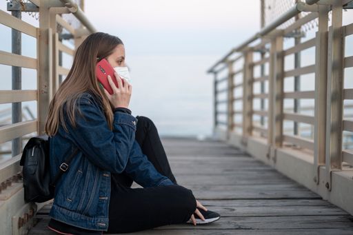 A stylish young Spanish woman wearing a facemask and talking on the phone while sitting on a bridge