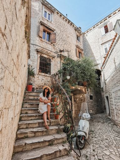 Lifestyle image of young woman wearing white summer dress sitting on stairs of picturesque house in old stone paved street with vintage vespa moped.